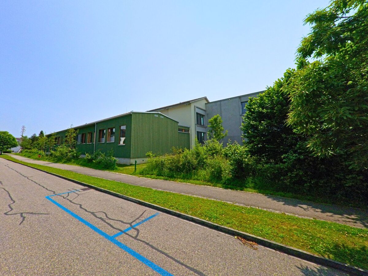 In Luterbach (4542, Kanton Solothurn) führt die Straße an einer gewerblich genutzten Fläche entlang, mit einem grünen Gebäude und Bäumen, die das Bild säumen.