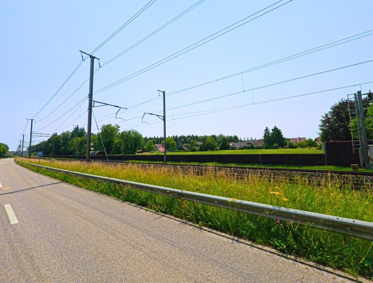 In Luterbach (4542, Kanton Solothurn) verläuft eine Strasse entlang der Eisenbahnlinie, die von Strommasten gesäumt wird und von grünen Wiesen flankiert ist.