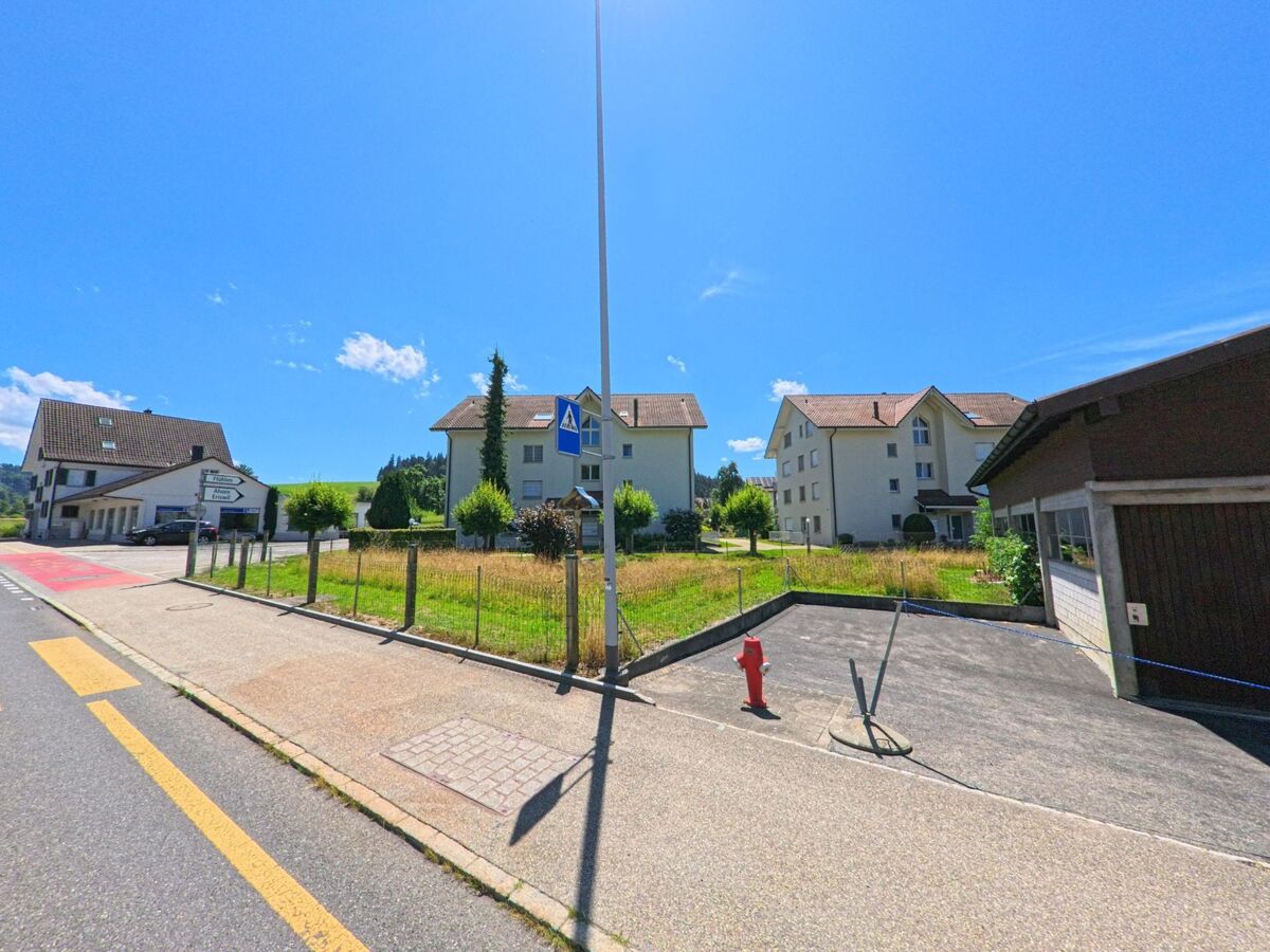 In Luthern (6154, Kanton Luzern) verläuft ein Trottoir mit Zebrastreifen entlang mehrerer Mehrfamilienhäuser und einem offenen Wiesengrundstück bei hellem Sonnenschein.