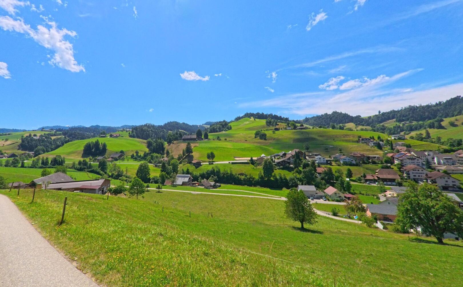 Eine gewundene Strasse führt oberhalb von Luthern (6156, Kanton Luzern) durch grüne Wiesenlandschaften mit Blick auf das weitläufige Dorf inmitten sanfter Hügel.