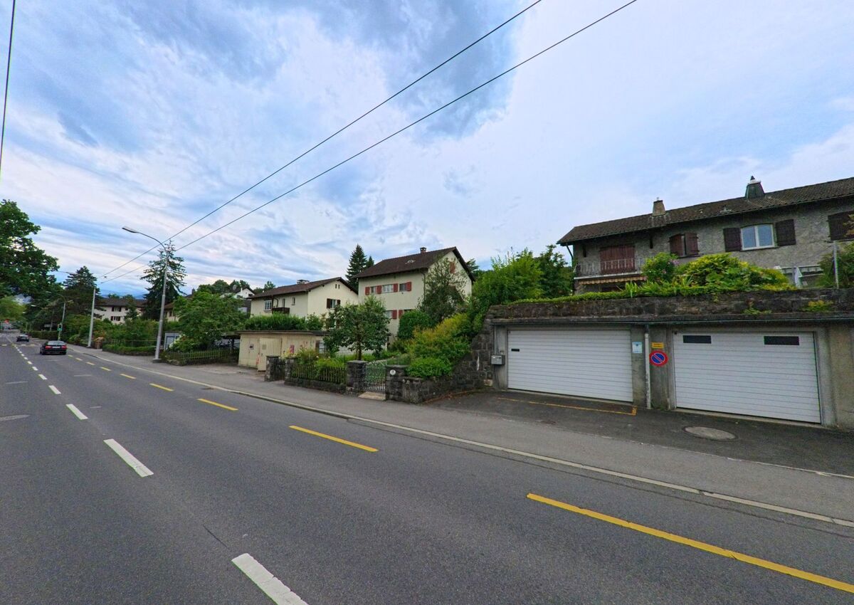 In Luzern (6005, Kanton Luzern) verlaufen Einfamilienhaeuser mit gepflegten Gaerten entlang einer ruhig gelegenen Strasse mit Fahrbahnmarkierungen und Laternen.