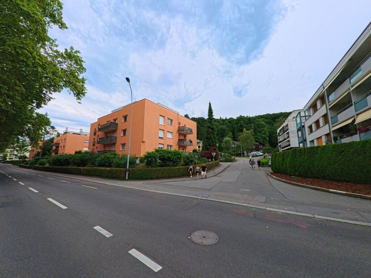 In einem Wohnquartier von Luzern (6005, Kanton Luzern) fuehren Gehwege zwischen modernen Mehrfamilienhaeusern mit orangefarbener Fassade und dichter Begruenung in Richtung Waldrand.