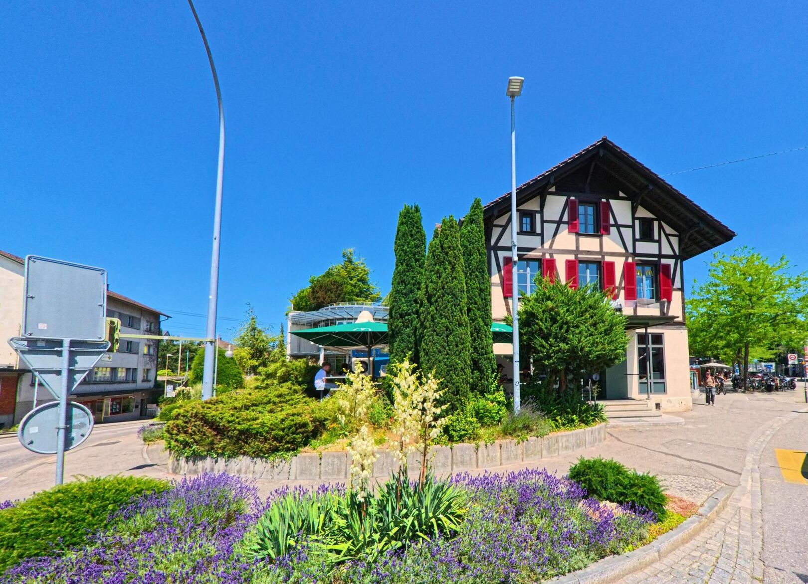Direkt beim Bahnhof von Lyss (3250, Kanton Bern) steht ein Fachwerkhaus mit roten Fensterläden hinter einem bunt bepflanzten Kreisel mit Lavendel und Yucca.