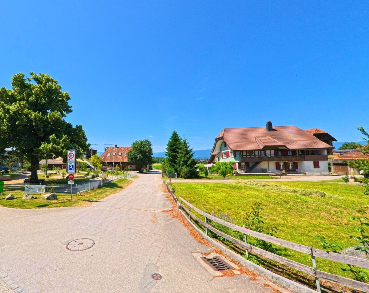 In Lyss (PLZ 3292, Kanton Bern) führt eine kleine Strasse an einem traditionellen Wohnhaus und einem Spielplatz mit schattigem Baumbestand vorbei.