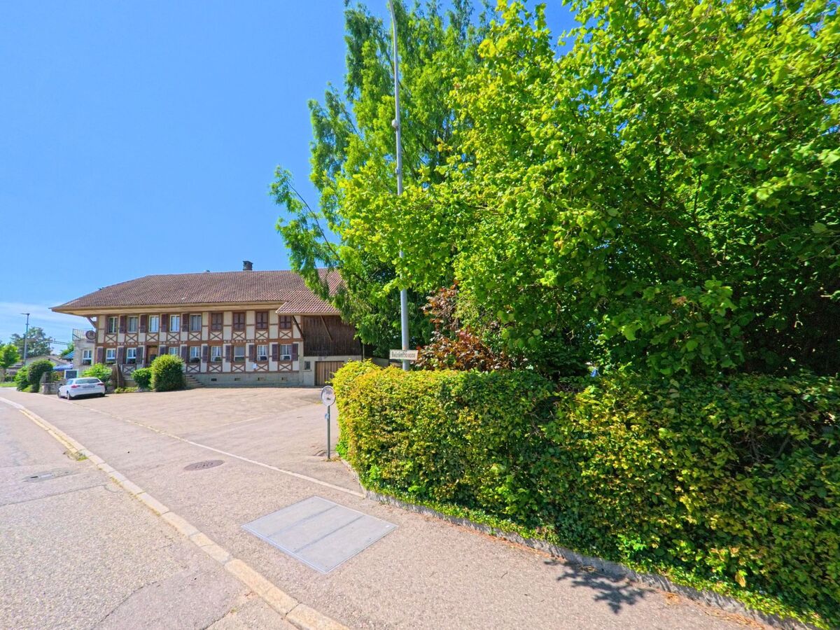 In Lyss (3292, Kanton Bern) steht ein langgezogenes, traditionelles Gebäude mit Fensterläden an einer ruhigen Strasse neben dichter Begrünung.