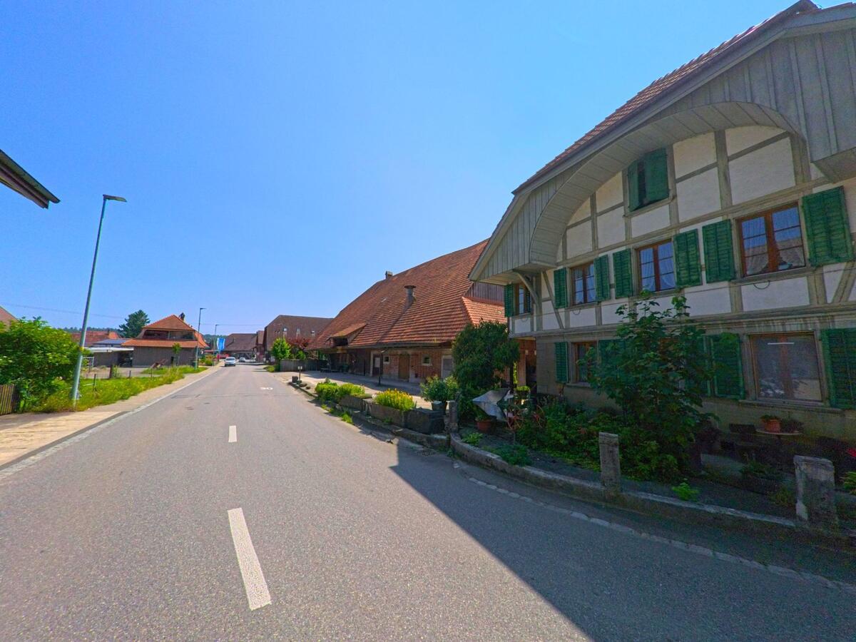 n Lyssach (3421, Kanton Bern) säumen traditionelle Bauernhäuser mit grünen Fensterläden eine zentrale Dorfstrasse mit Vorgärten und ruhigem Verkehr.
