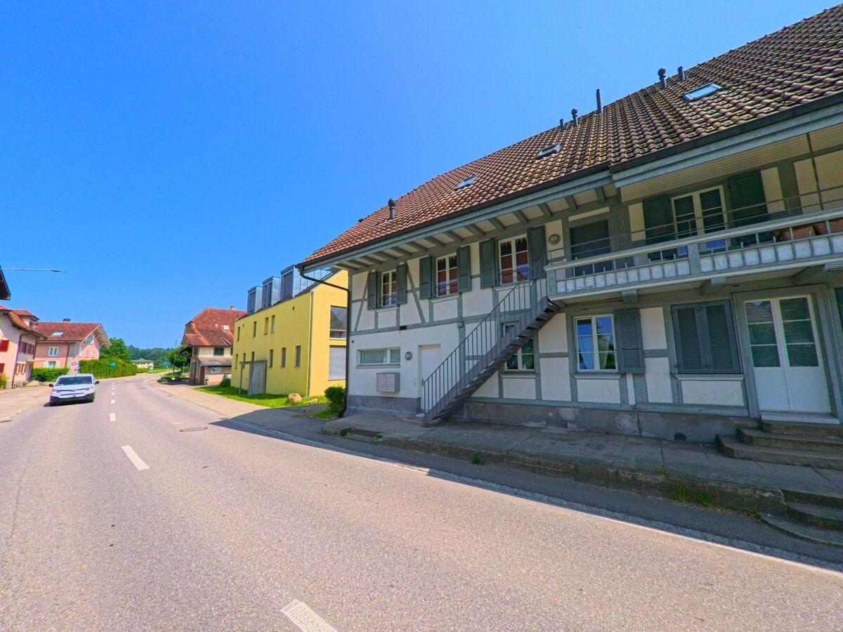 In Lyssach (PLZ 3421, Kanton Bern) säumen traditionelle und moderne Wohnhäuser eine verkehrsberuhigte Dorfstrasse mit Sicht auf angrenzende Felder.