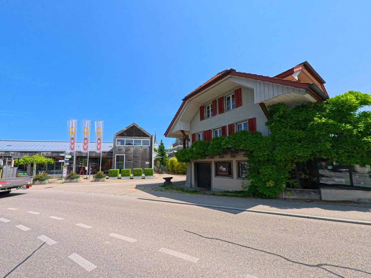 Im Zentrum von Madiswil (4934, Kanton Bern) steht ein mit Pflanzen bewachsenes Haus neben einem modernen Coop-Gebäude mit Glasfassade und auffälligen Flaggen.
