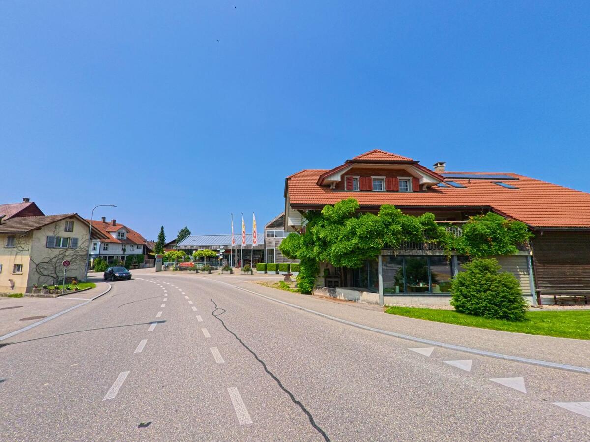 Eine sanft geschwungene Strasse führt in Madiswil (4934, Kanton Bern) am bewachsenen Gebäude mit rotem Ziegeldach vorbei in Richtung Coop und weiteren Wohnhäusern.