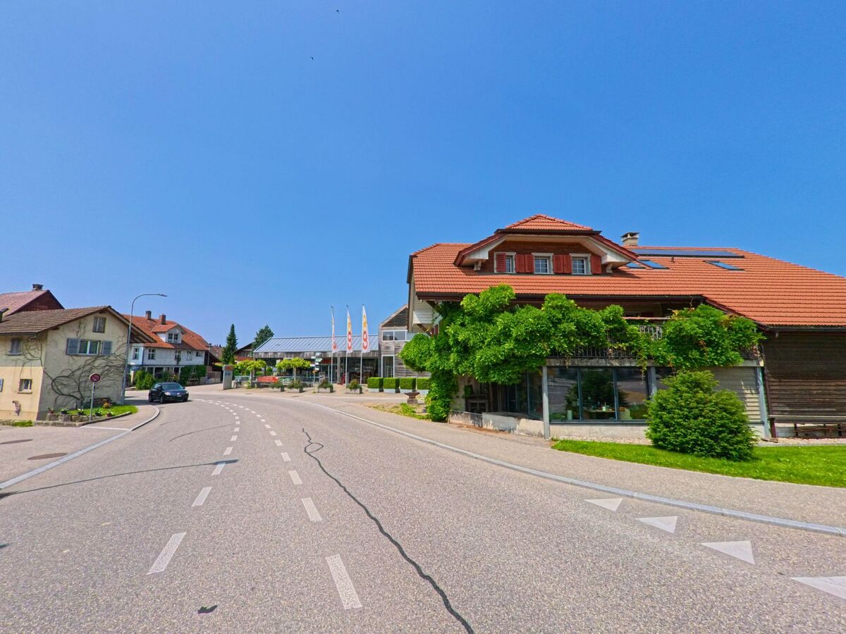 Eine sanft geschwungene Strasse führt in Madiswil (4934, Kanton Bern) am bewachsenen Gebäude mit rotem Ziegeldach vorbei in Richtung Coop und weiteren Wohnhäusern.