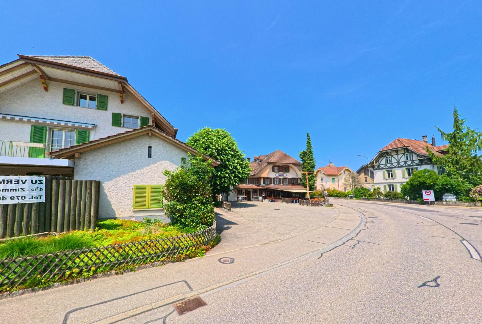 In Madiswil (4934, Kanton Bern) säumen gepflegte Häuser mit charakteristischen Fassaden eine leicht geschwungene Dorfstrasse unter klarem Sommerhimmel.