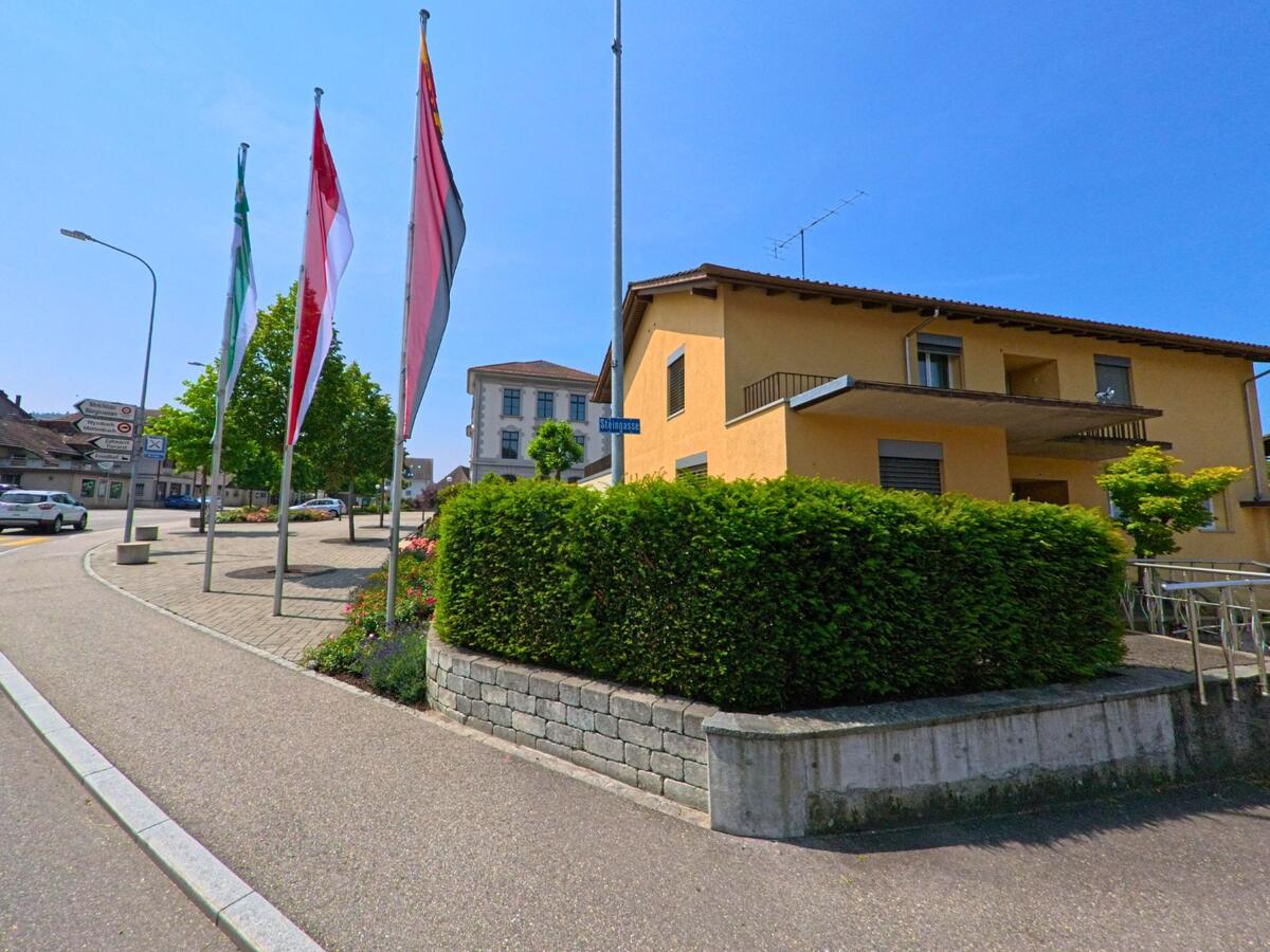 In Madiswil (4934, Kanton Bern) säumen Fahnenmasten mit Gemeindeflaggen einen breiten Gehweg vor einem gelben Wohnhaus an der Striggasse.