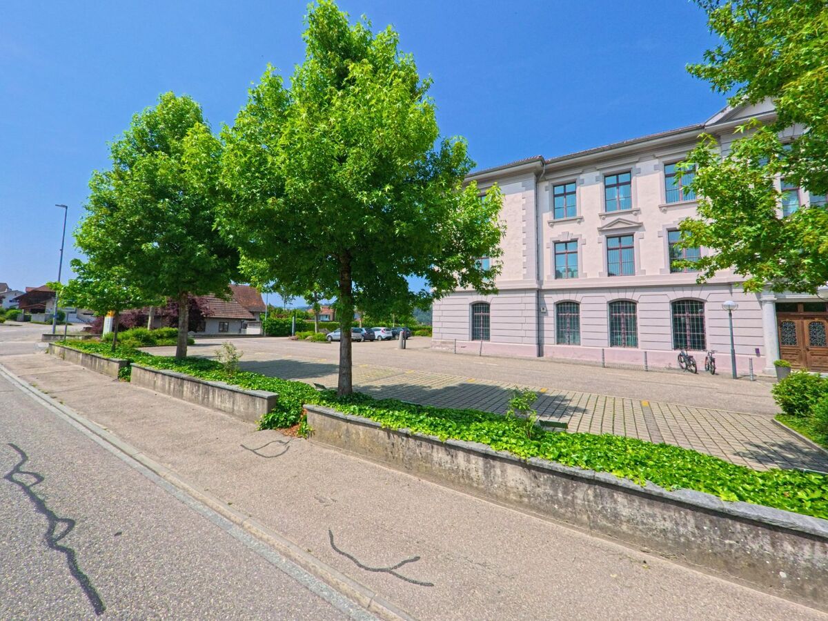 Vor einem historischen Gebäude in Madiswil (4934, Kanton Bern) erstreckt sich ein baumbestandener Parkplatz mit angrenzendem Gehweg und gepflegtem Grünstreifen.