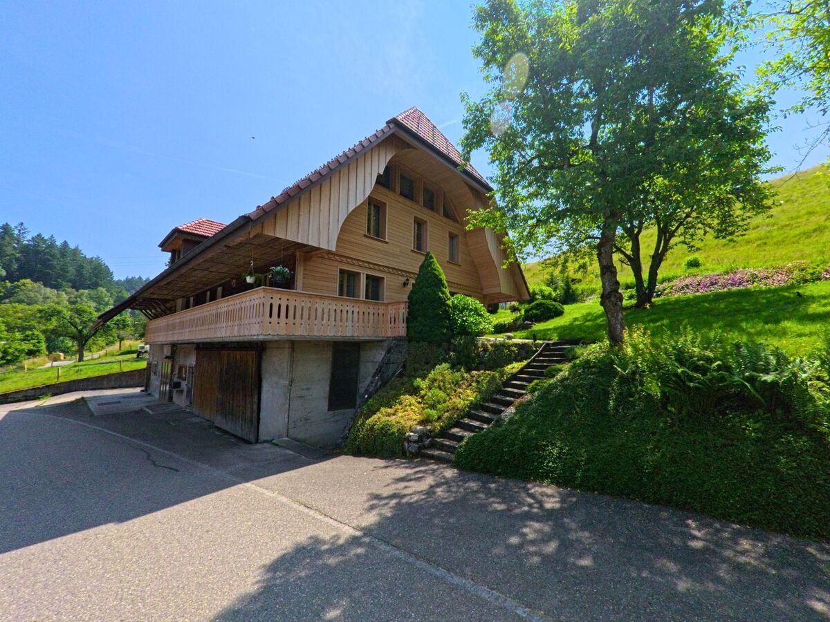 Ein traditionelles Holzhaus mit Balkon steht in Madiswil (4934, Kanton Bern) am Hang, umgeben von gepflegtem Grün und einer Treppe, die durch den Garten nach oben führt.