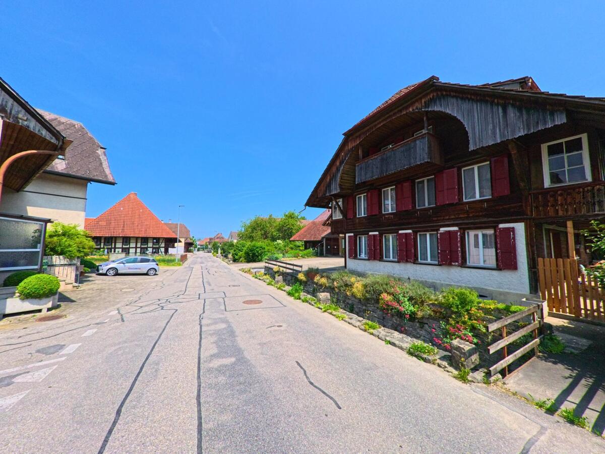 Entlang einer ruhigen Quartierstrasse in Madiswil (4934, Kanton Bern) reihen sich traditionelle Bauernhäuser mit roten Fensterläden und gepflegten Vorgärten aneinander.
