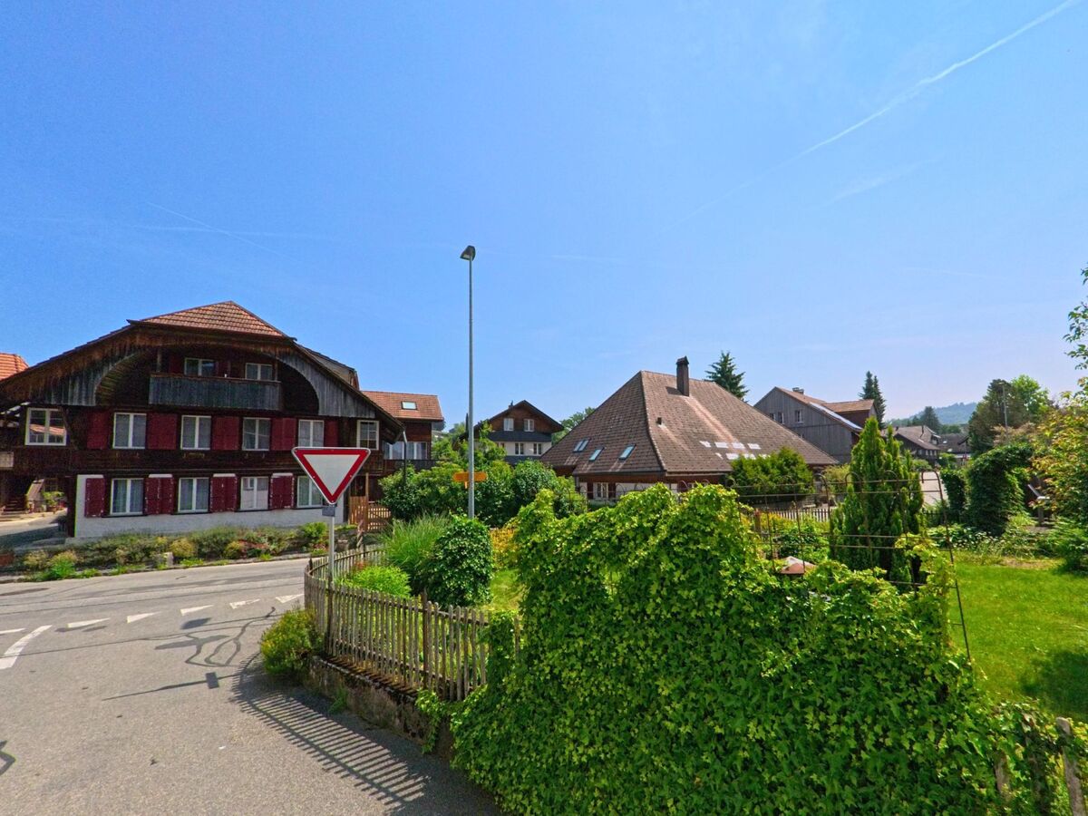 In Madiswil (4934, Kanton Bern) treffen sich zwei Strassen vor einem alten Haus mit markantem Balkon und roten Fensterläden, flankiert von grünen Gärten und einem Verkehrsschild.