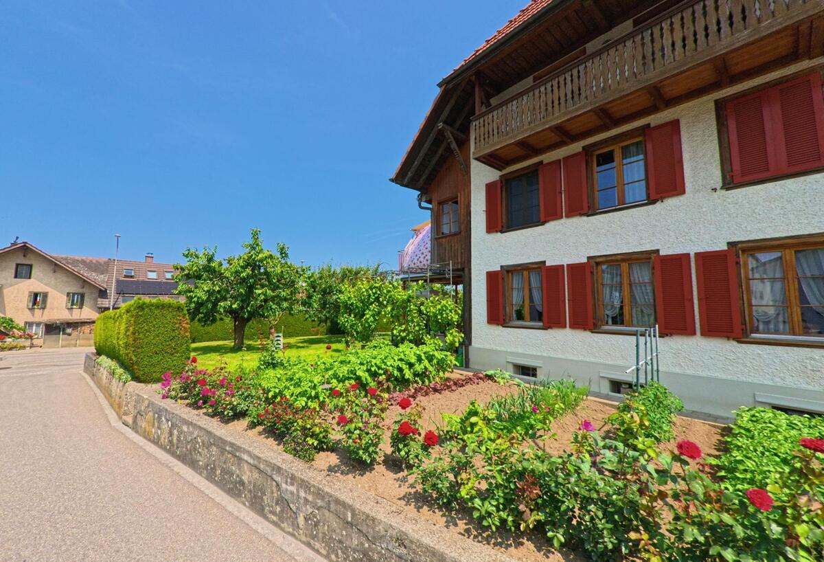 In einem Vorgarten in Madiswil (4934, Kanton Bern) blühen rote Rosen und andere Pflanzen vor einem gepflegten Haus mit roten Fensterläden und hölzernem Balkon.