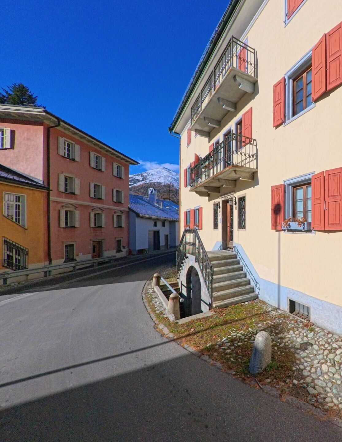 Zwischen farbigen Wohnhäusern mit Balkonen verläuft eine ruhige Dorfstrasse in Madulain (7523, Kanton Graubünden), im Hintergrund erhebt sich ein schneebedeckter Gipfel.