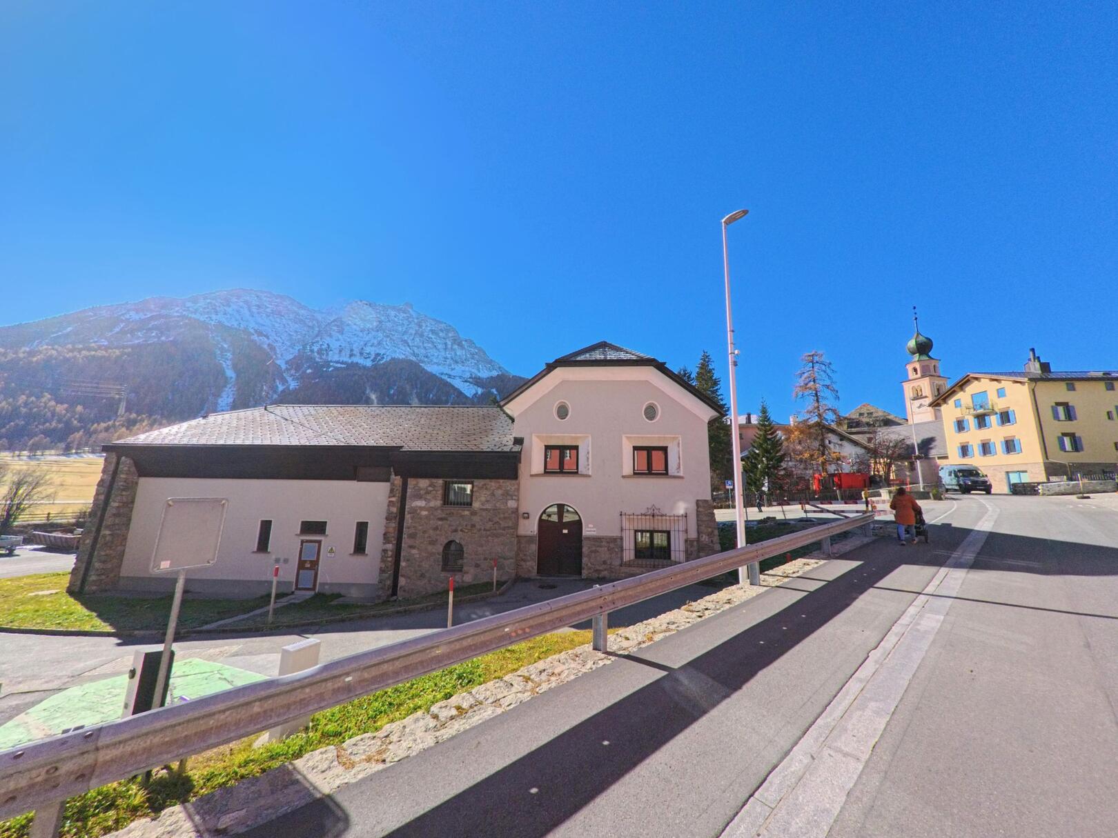 Entlang einer leicht ansteigenden Strasse mit Blick auf historische Gebäude zeigt sich ein Teil des Dorfkerns von Madulain (7523, Kanton Graubünden) vor alpiner Bergkulisse.