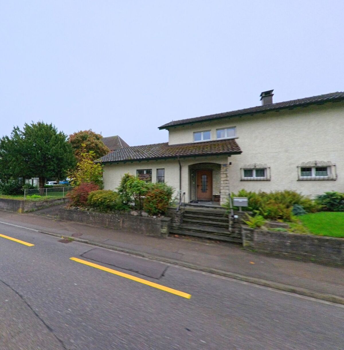 Ein Einfamilienhaus mit Vorgarten und gedecktem Eingang steht an einer Hauptstrasse in Mägenwil (5506, Kanton Aargau) zwischen Bäumen und Sträuchern.