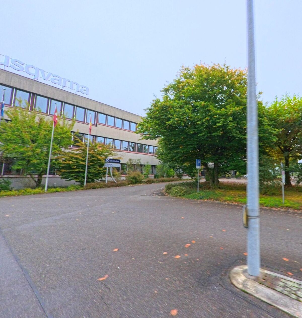 Das Firmengebäude von Husqvarna in Mägenwil (5506, Kanton Aargau) ist von Bäumen und Flaggen gesäumt und liegt an einer Zufahrtsstrasse mit Parkplatz.