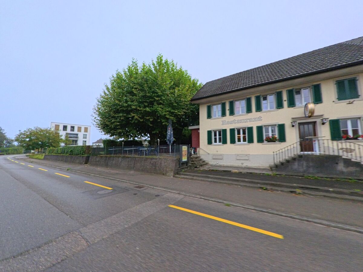 Ein traditionelles Restaurant mit grünen Fensterläden steht an einer leicht gebogenen Strasse in Mägenwil (5506, Kanton Aargau), daneben befindet sich eine Terrasse unter grossen Bäumen.