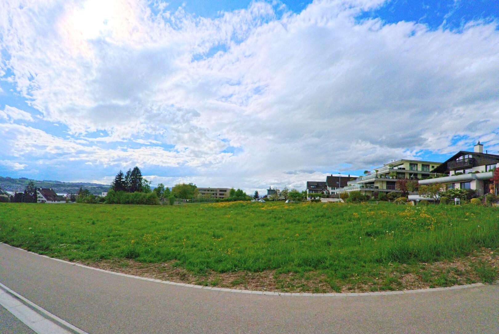 In Männedorf im PLZ-Gebiet 8708 im Kanton Zürich öffnet sich ein weiter Blick über eine grüne Wiese mit modernen Wohnhäusern im Hintergrund und einem teils bewölkten Himmel darüber.
