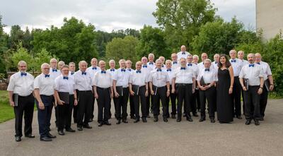 Der Männerchor steht in weissen Hemden mit blauen Fliegen in Reinach (4153, Kanton Baselland) geschlossen im Freien und haelt Notenmappen in den Haenden.