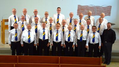 Der Männerchor Sins (5643, Kanton Aargau) steht in weissen Hemden und blauen Krawatten auf einer Kirchenbühne zu einem Gruppenfoto versammelt.