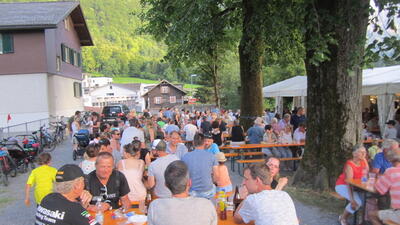 Beim Anlass des Männerturnvereins MTV Mollis in Glarus Nord (8753, Kanton Glarus) sitzen zahlreiche Personen an langen Tischen unter Bäumen und Zelten zusammen.