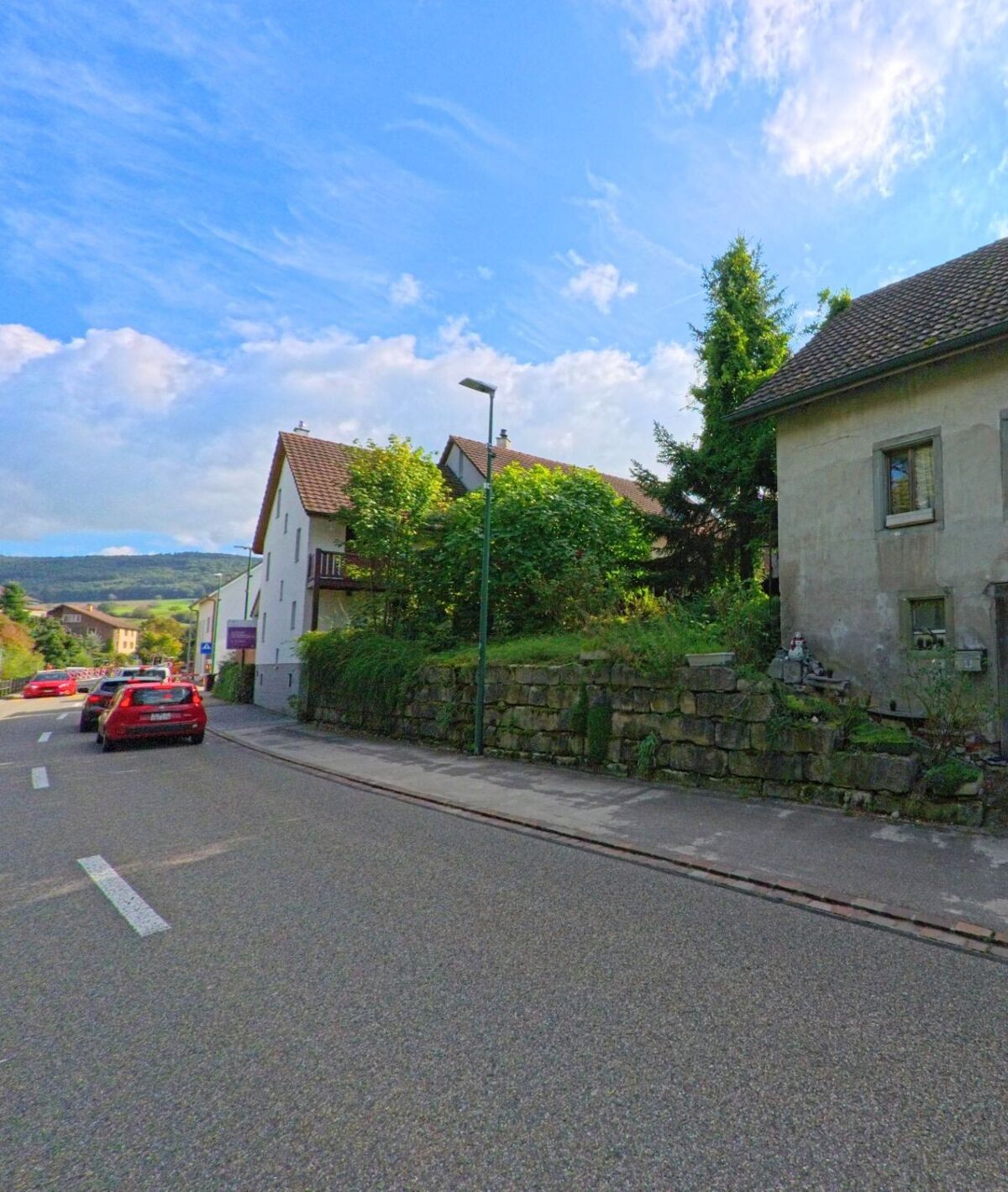 An einer leicht ansteigenden Strasse in Magden (4312, Kanton Aargau) stehen Wohnhäuser mit Gärten und Natursteinmauern, im Hintergrund sind grüne Hügel zu erkennen.