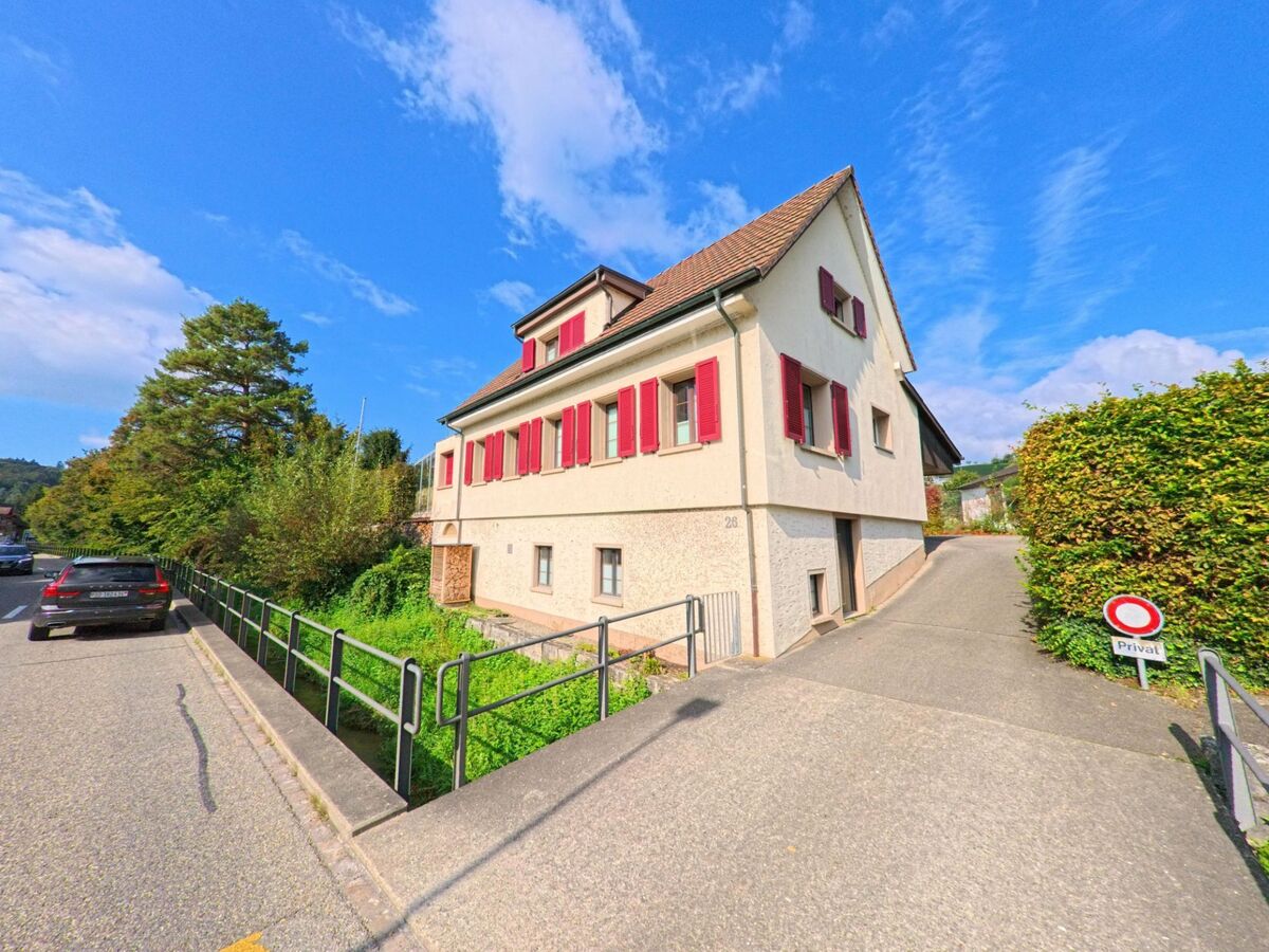 Ein helles Wohnhaus mit roten Fensterläden steht an einer Nebenstrasse in Magden (4312, Kanton Aargau), daneben führt ein schmaler Weg an einer Hecke entlang.