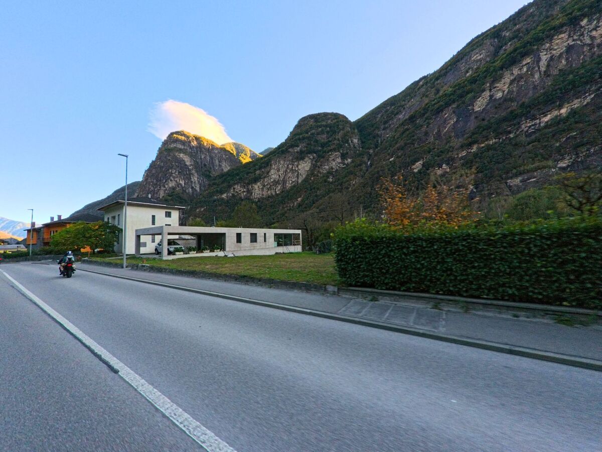 Der Kirchturm von Maggia (6674, Kanton Tessin) erhebt sich vor den sonnenbeschienenen Hängen des Maggiatals, umgeben von traditionellen Häusern und grünen Wiesen.