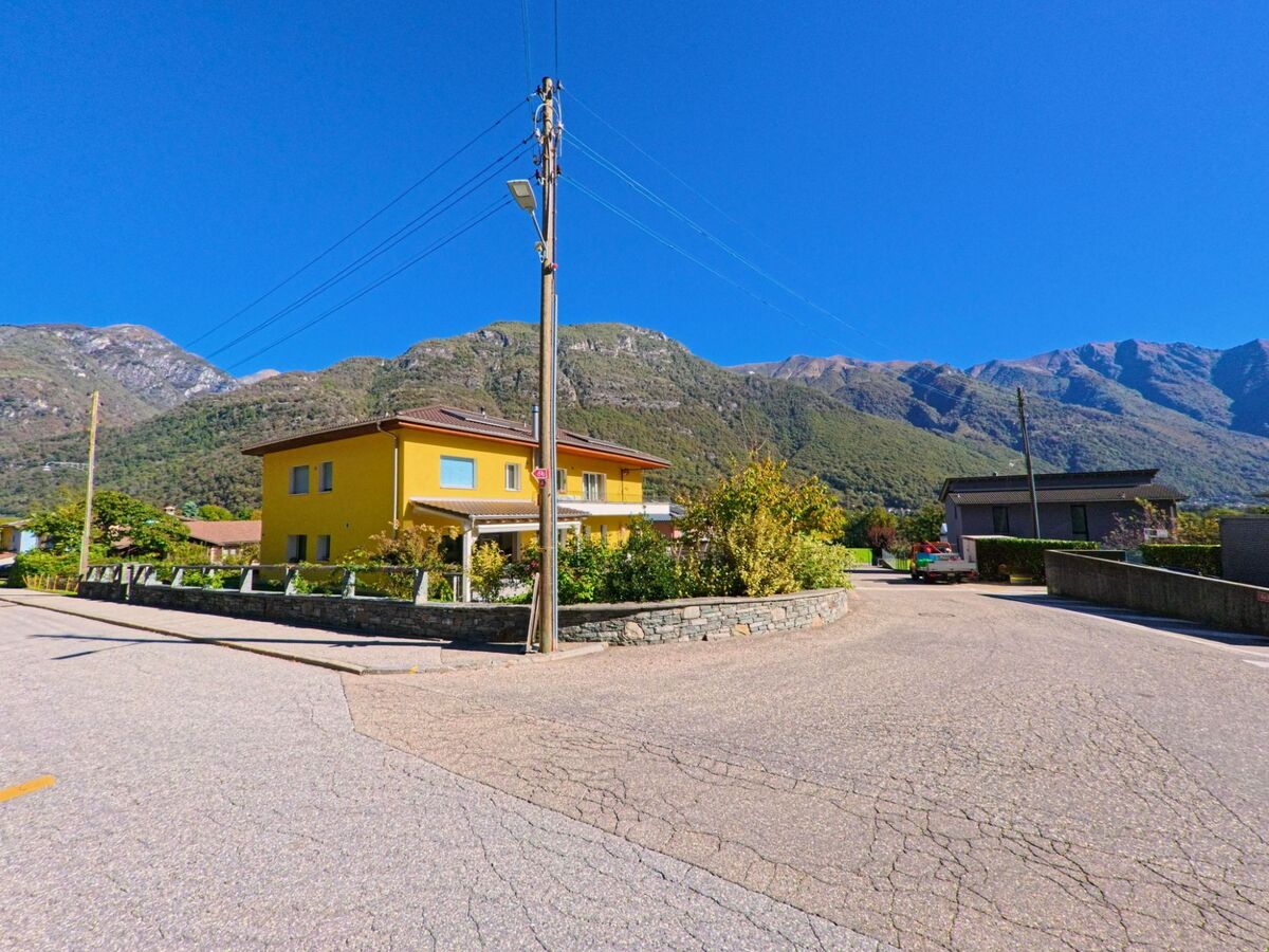 Ein gelbes Wohnhaus steht in Maggia (6677, Kanton Tessin) an einer ruhigen Strasse mit Blick auf die bewaldeten Hänge der umliegenden Alpen.