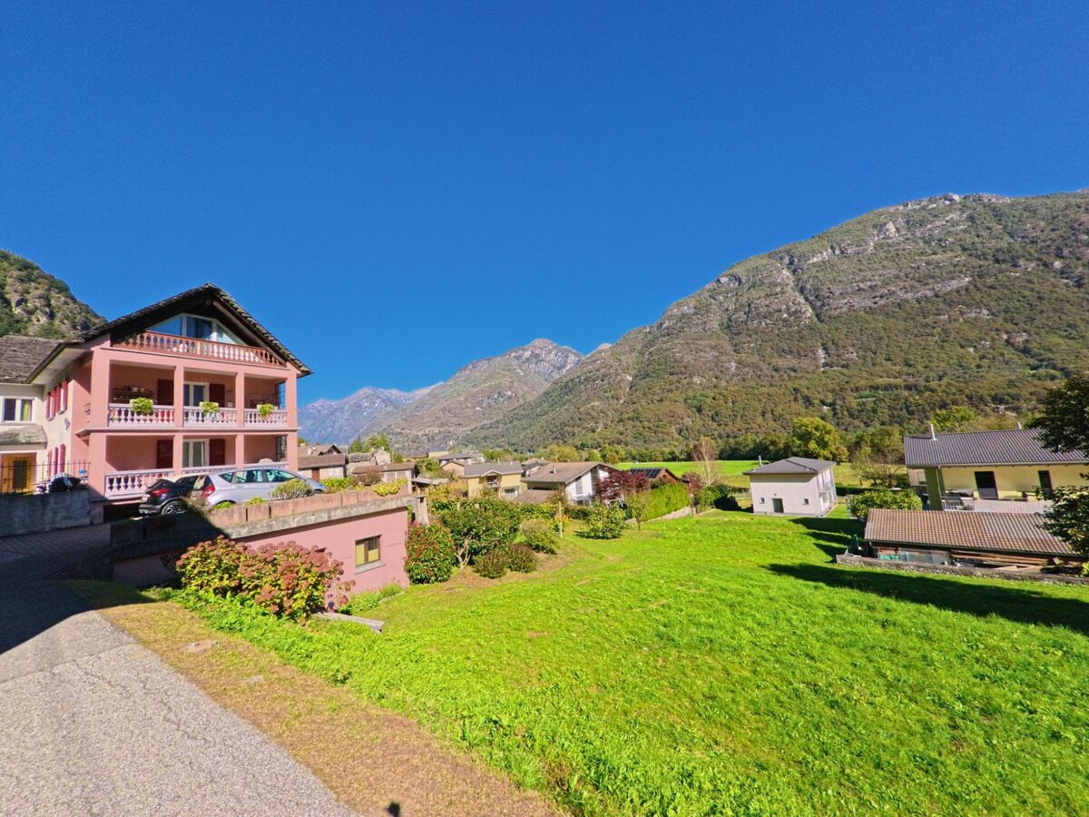 Ein rosafarbenes Wohnhaus mit Balkonen steht in Maggia (6677, Kanton Tessin) vor einer beeindruckenden Bergkulisse, umgeben von grünen Wiesen und klarer Alpenluft.