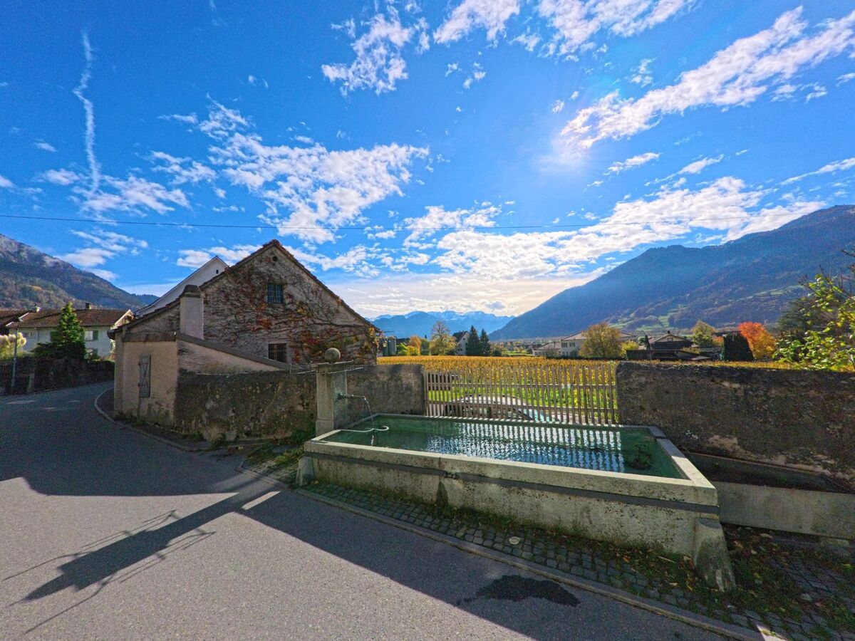 Neben einer schmalen Strasse liegt ein steinerner Dorfbrunnen vor einer ummauerten Wiesenfläche in Maienfeld (7304, Kanton Graubünden), mit weiter Sicht auf das Rheintal und die umliegenden Berge.