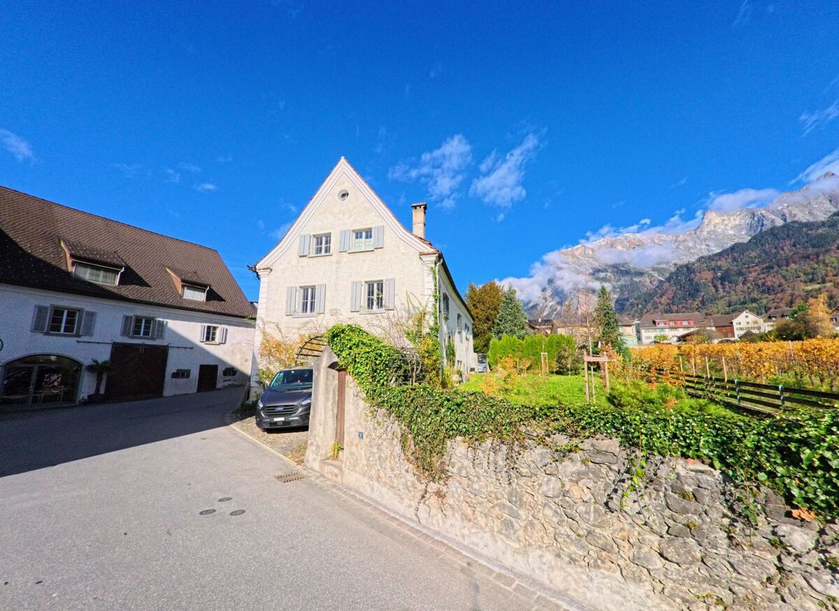 Entlang einer schmalen Strasse steht ein freistehendes Wohnhaus mit Gartenmauer in Maienfeld (7304, Kanton Graubünden), dahinter öffnen sich Rebflächen und ein alpines Bergpanorama.