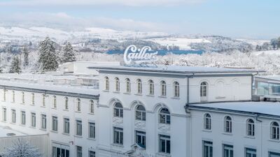 In Broc (1636, Kanton Freiburg) erhebt sich das schneebedeckte Gebaeude der Maison Cailler vor einer winterlichen Landschaft mit verschneiten Baeumen und huegeligem Hintergrund.