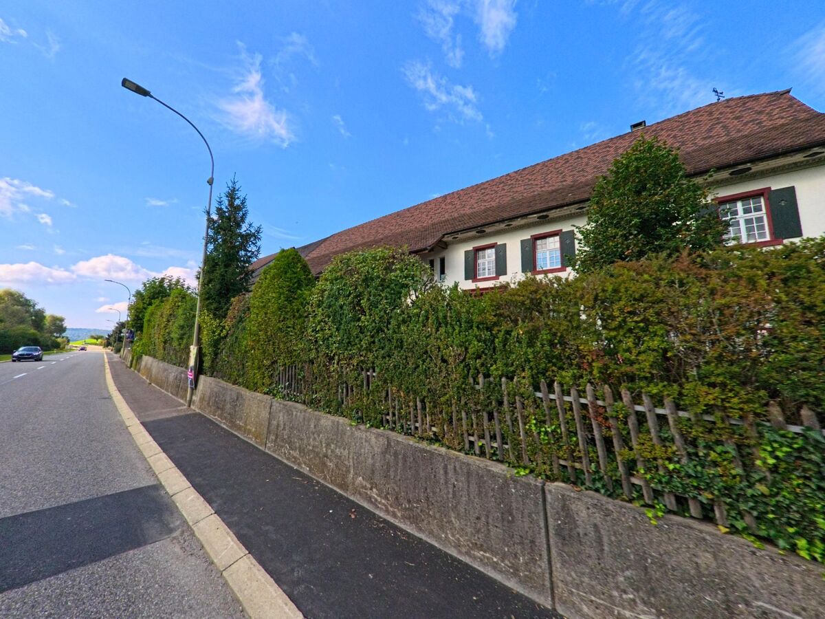 Entlang der Strasse in Maisprach (4464, Kanton Basel-Landschaft) verläuft eine dichte Hecke vor einem traditionellen Haus mit grünen Fensterläden und rotem Dach.