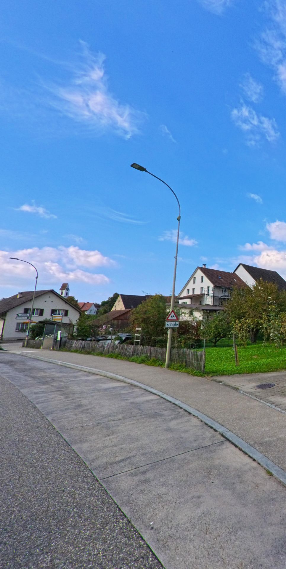 Ein Schulhinweisschild steht am leicht ansteigenden Strassenrand in Maisprach (4464, Kanton Basel-Landschaft), während dahinter Wohnhäuser und Gärten vor dem blauen Himmel sichtbar sind.