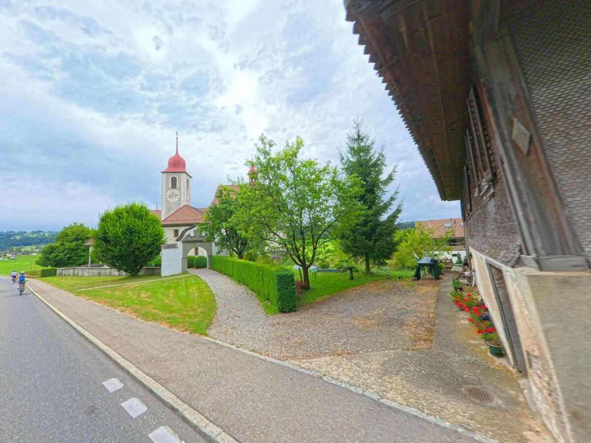In Malters (6002, Kanton Luzern) liegt eine Kirche mit rotem Zwiebelturm und Uhr am Rande einer Landstrasse, flankiert von gepflegten Grünflächen und alten Wohnhäusern.