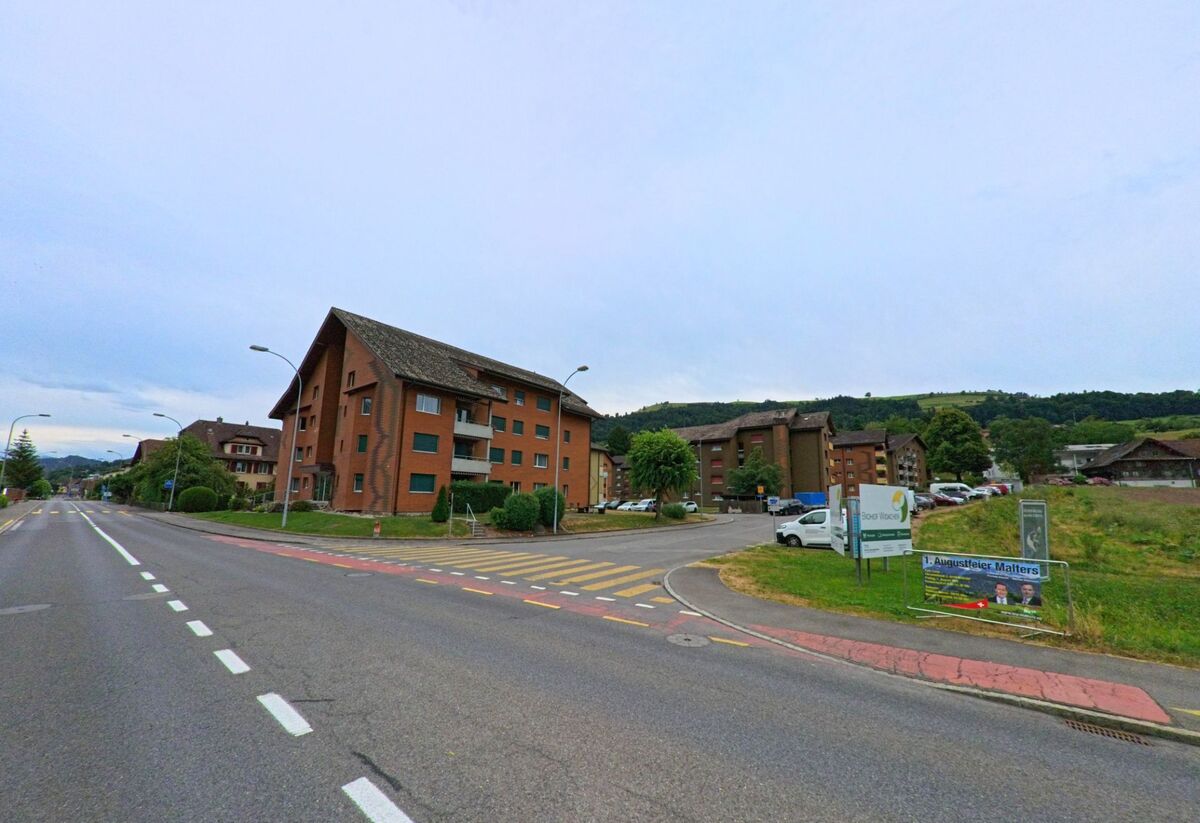 In Malters (6002, Kanton Luzern) führt eine breite Strasse an einem Wohnquartier mit roten Mehrfamilienhäusern vorbei, auf einer Wiese steht ein Plakat für eine Veranstaltung.