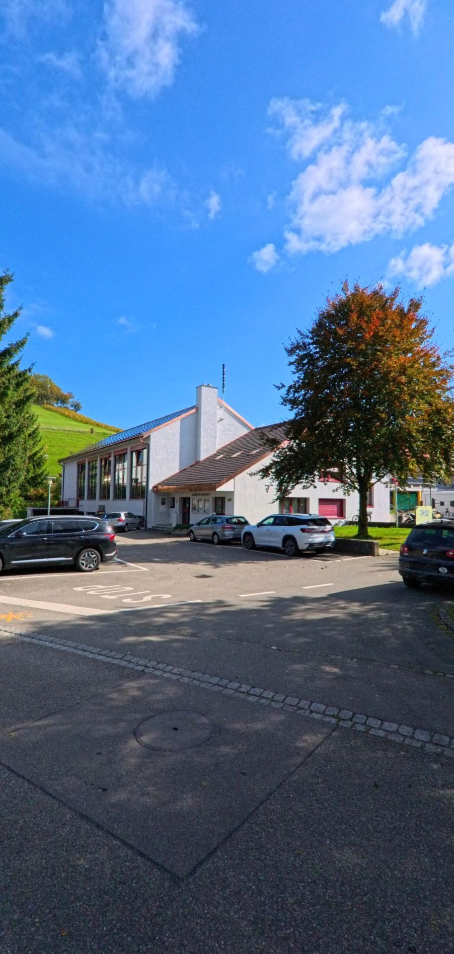 Vor dem Gemeindehaus von Mandach (5318, Kanton Aargau) stehen mehrere geparkte Autos, während sich im Hintergrund grüne Hügel und Bäume erheben.