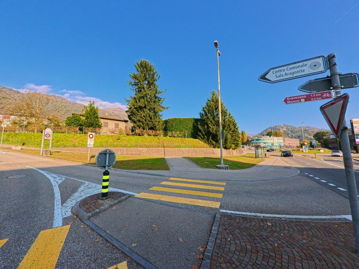 An einem Fussgängerstreifen mit Wegweisern öffnet sich eine Kreuzung in Manno (6928, Kanton Tessin), umgeben von Grünflächen, Bäumen und sanft ansteigender Hügellandschaft.