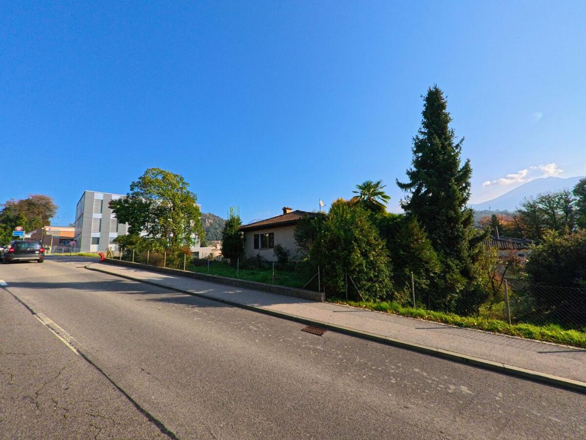 Eine ruhige Wohnstrasse mit Einfamilienhäusern und dichter Bepflanzung verläuft durch Manno (6928, Kanton Tessin) unter klarem Himmel und mit Blick auf die umliegende Hügellandschaft.