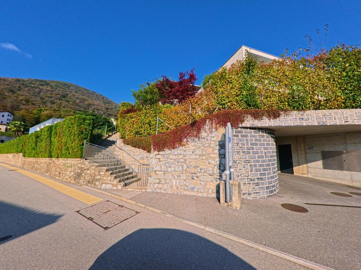 Entlang einer ruhigen Nebenstrasse führen Steintreppen zu terrassierten Gärten und Wohnhäusern in Manno (6928, Kanton Tessin), eingebettet in eine begrünte Hanglage.