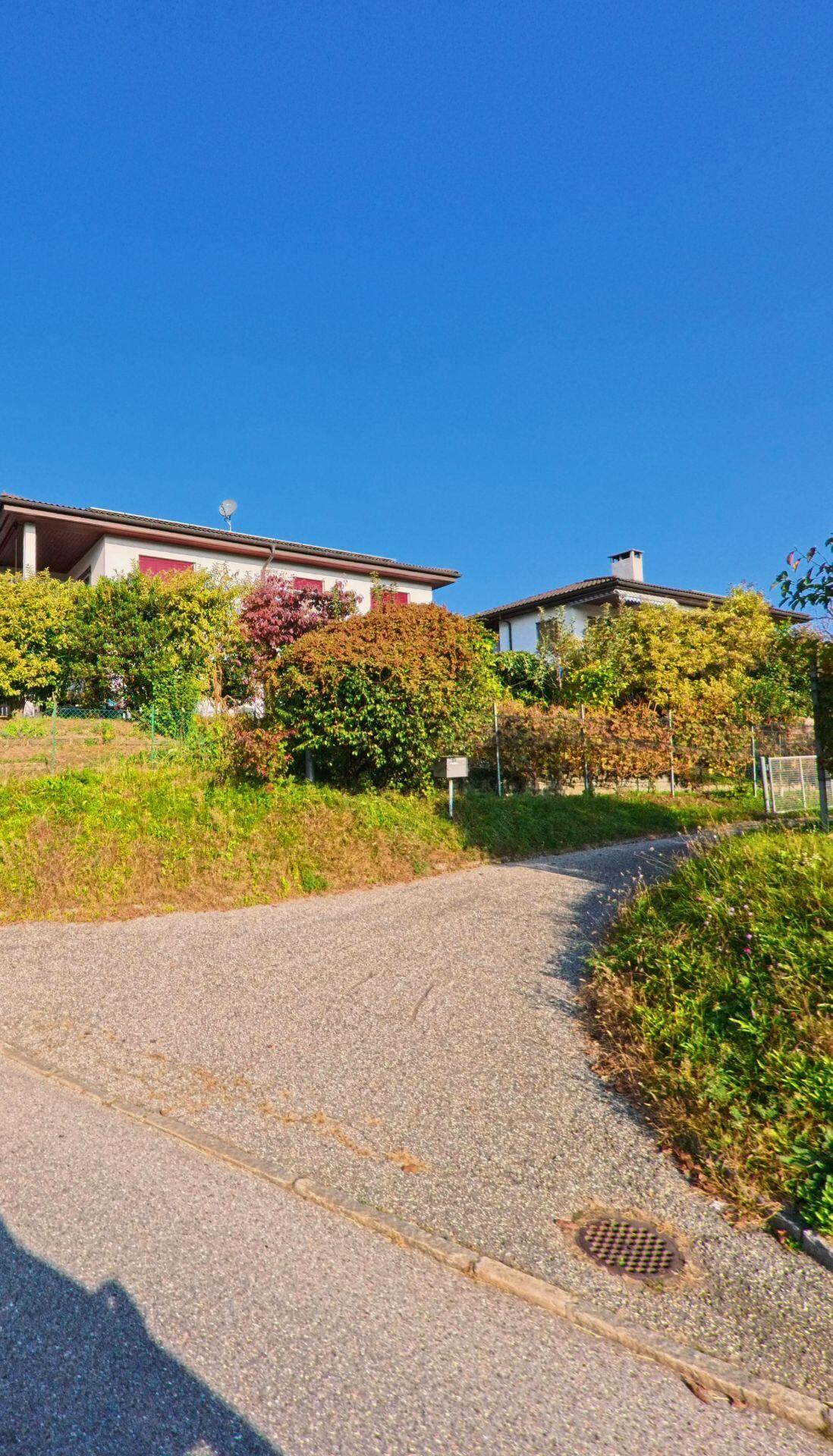 Ein schmaler Zufahrtsweg führt zu Wohnhäusern mit Gärten in Manno (6928, Kanton Tessin), umgeben von herbstlicher Vegetation unter klarem Himmel.