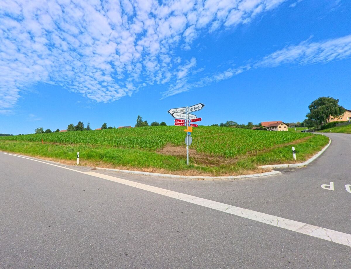 Auf einem Strassenabzweiger in Maracon (1613, Kanton Waadt) stehen Wegweiser vor einem weiten Feld mit Blickrichtung Palézieux und Semsales unter klarem Himmel.