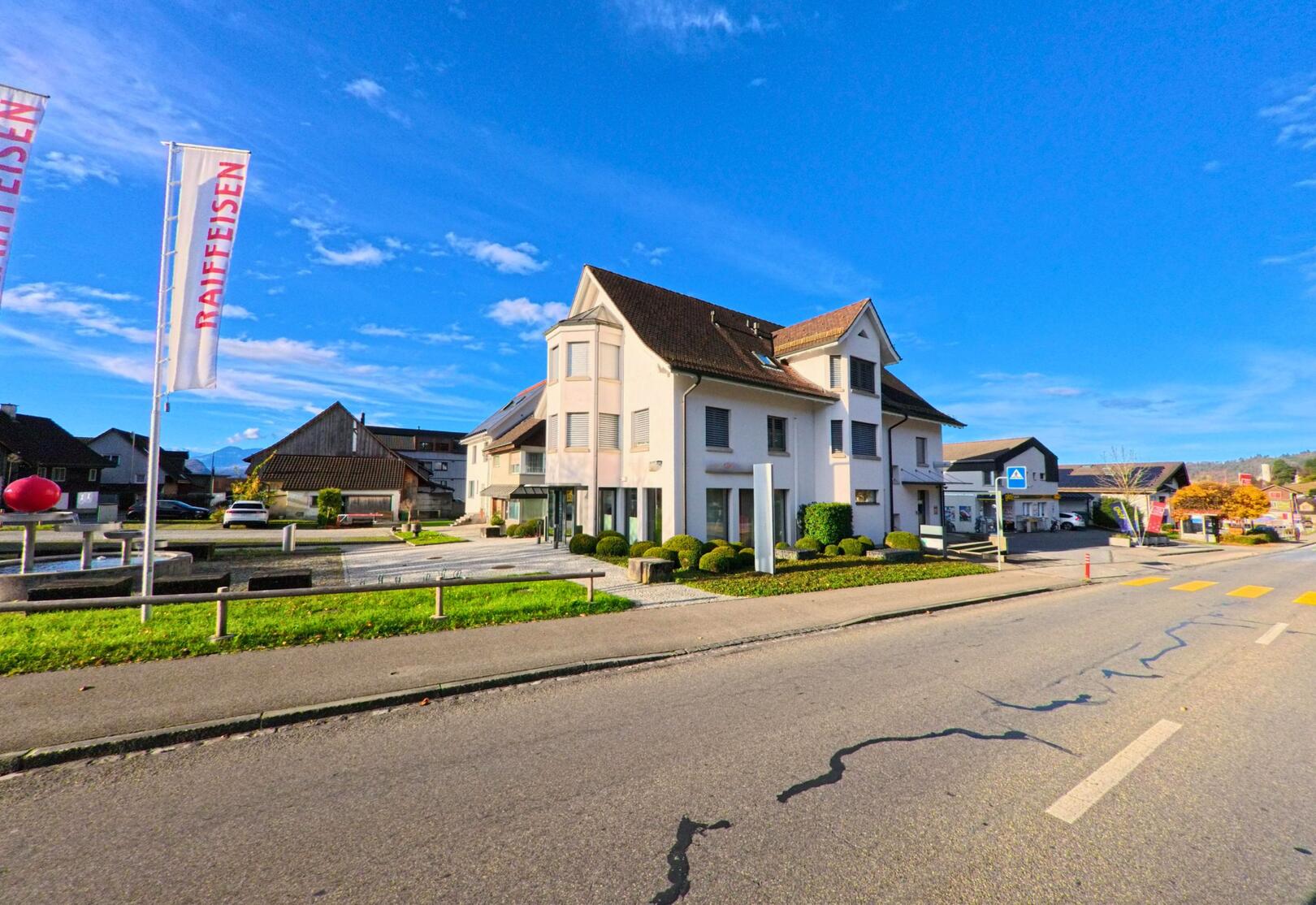 Vor dem hellen Bankgebaeude in Marbach (9437, Kanton St. Gallen) stehen Raiffeisen-Fahnen an einer breiten Strasse mit Zebrastreifen unter klarem Himmel.