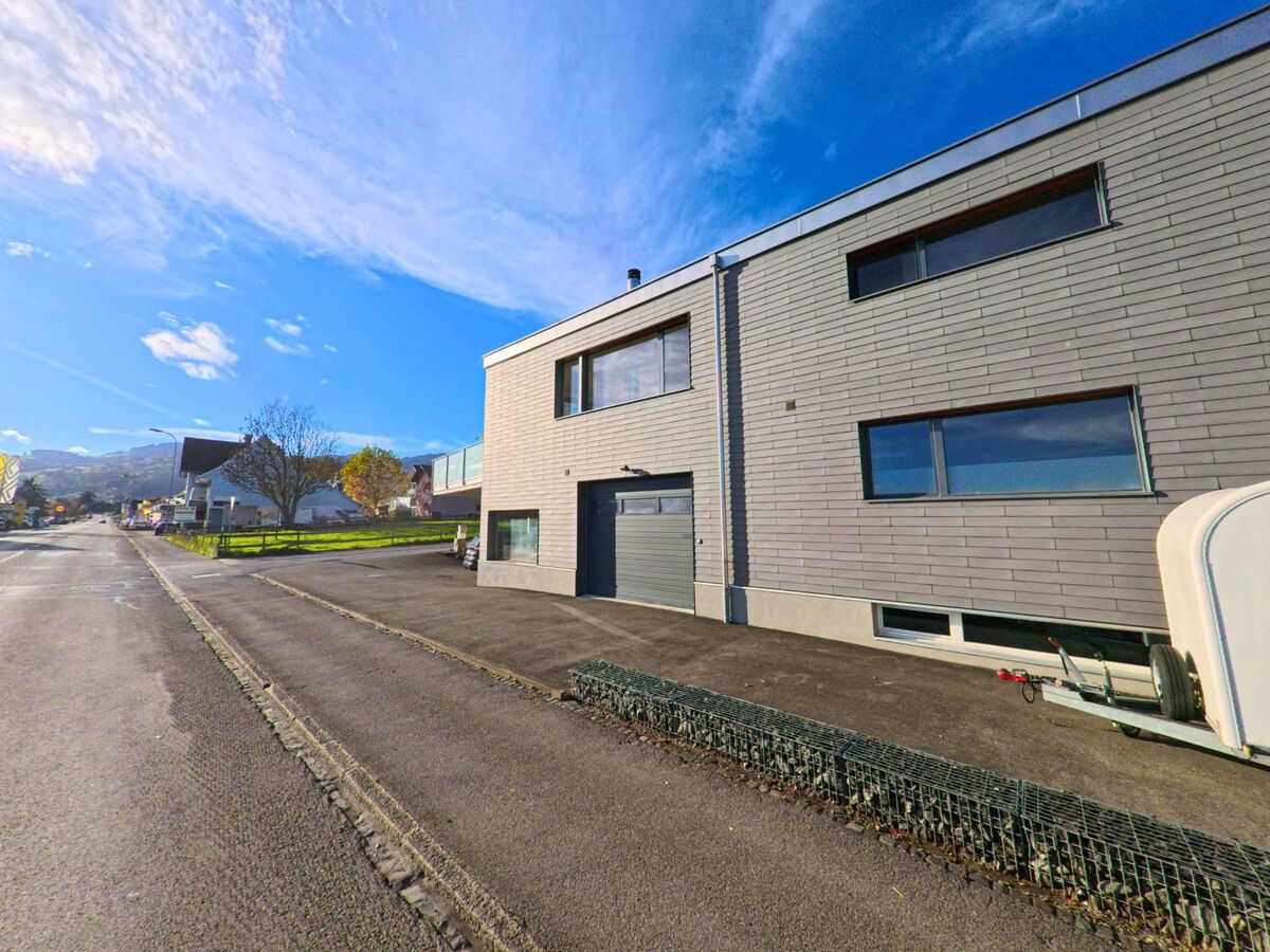 Ein modernes Wohngebaeude mit klarer Linienfuehrung befindet sich in Marbach (9437, Kanton St. Gallen) direkt an einer breiten Strasse mit Blick auf die umliegenden Huegel.
